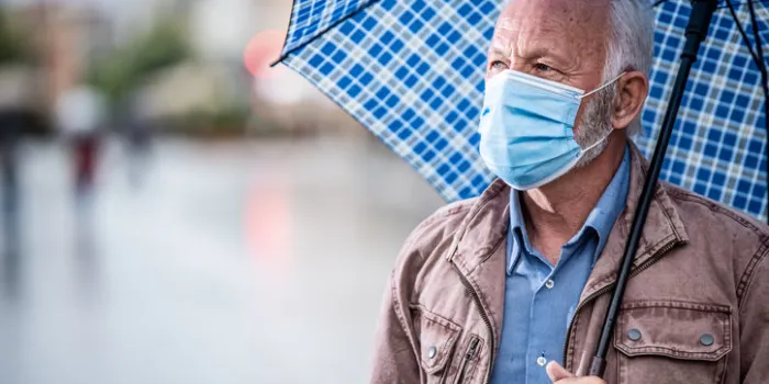 photo of senior man walking in the city on rainy day under umbrella wearing protective face mask