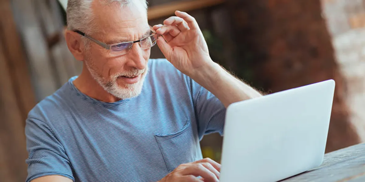 surf the internet waist up of a cheerful aged man sitting at the table and using his laptop while touching his glasses