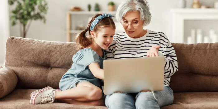 happy aged woman with cute granddaughter smiling while sitting on sofa and using laptop in living room