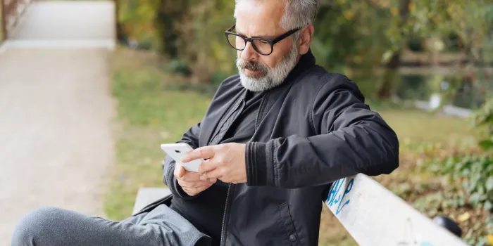 attractive mature bearded man wearing glasses relaxing on a park bench sending a text message on his mobile phone