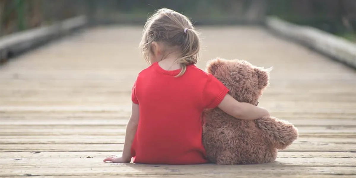little girl in a red dress sitting on a boardwalk hugging teddybear, view from behind