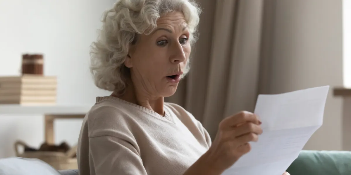 amazed shocked mature woman with wide open eyes and mouth reading letter close up, holding paper notification in hands, sitting on couch at home, overjoyed middle aged female received unexpected news