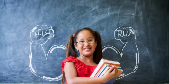concept on blackboard at school intelligent and successful hispanic girl in class portrait of female child smiling, looking at camera, holding books against drawing of muscles on blackboard