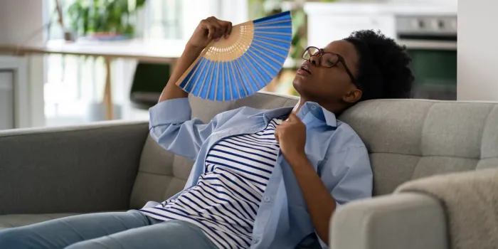 tired exhausted african american woman sits on sofa at home using paper fans and closing eyes suffering from stuffiness tormented black girl housewife is resting after hot walk in summer weather