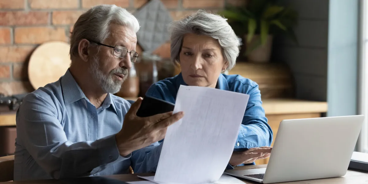 focused worried grey haired elderly spouses sit in kitchen with laptop on table reading financial papers documents, checking bills looking concerned, planning budget expenses, discussing debt concept