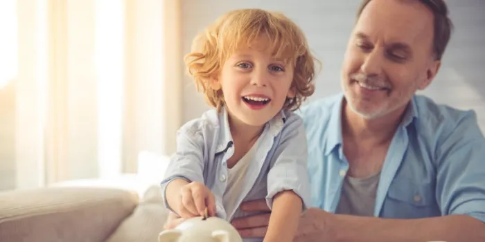 mignon petit garçon met la pièce dans la tirelire, regardant la caméra et souriant tout en passant du temps avec son grand-père à la maison