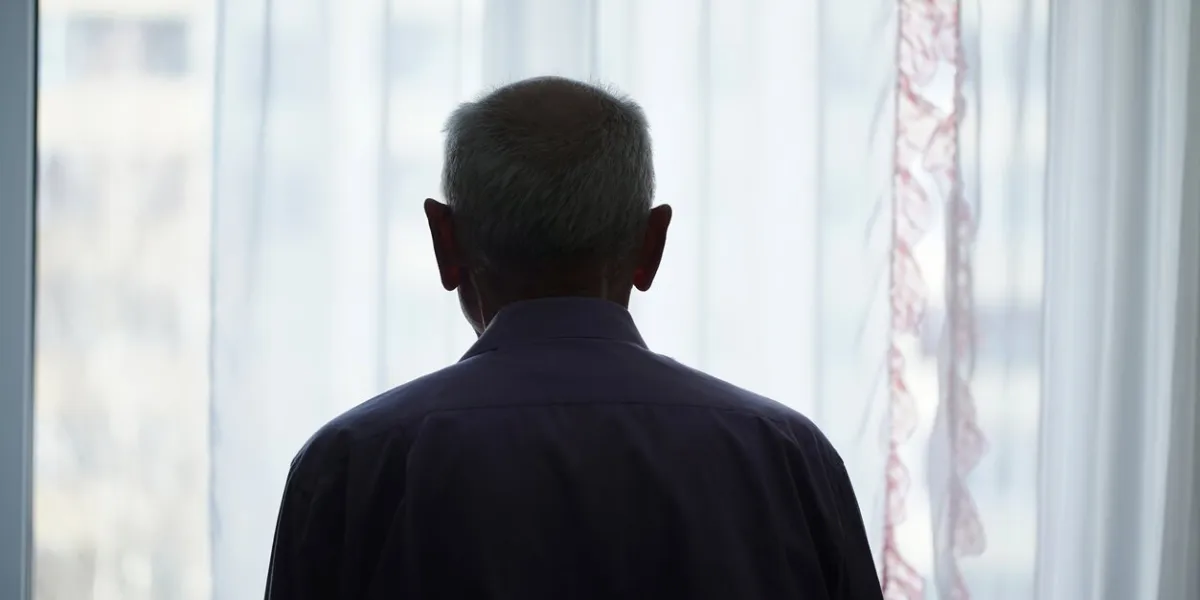 silhouette of retired man looking through window with transparent curtain standing at home rear view loneliness and old human care concept