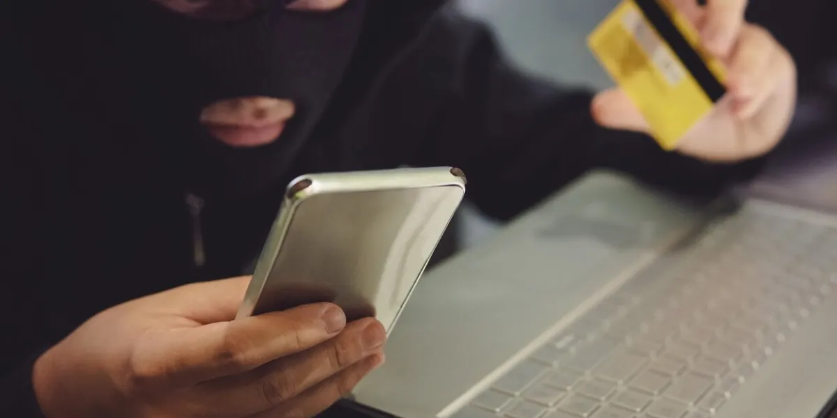 male hacker in a robber mask uses phone, credit card and laptop in some fraudulent scheme cyber thief stole the personal data and credit card information hacker uses malware to steal user's money