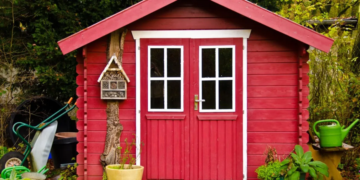 a light red small shed, gardenhouse, with some garden tools around it