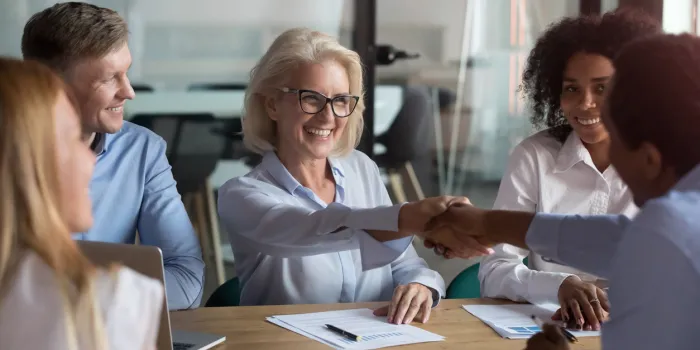 middle aged woman executive manager company representative welcoming business partner while sitting in modern office boardroom or trainer shake hands with participant before starting education meeting