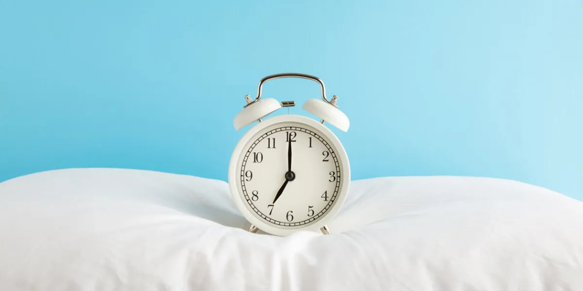 white alarm clock on pillow at light blue wall background pastel color 7 o'clock in morning waking time concept closeup front view