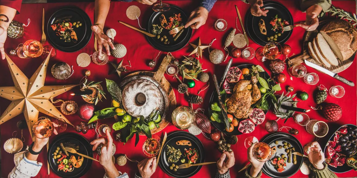friends celebrating christmas flat-lay of people eating and talking over festive table with red cloth with champagne, roasted chicken, bundt cake, fruits, decorations, top view winter holiday party