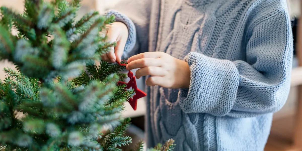 little child girl is decorating christmas tree with red ornaments kid is wearing in cotton knitted blue sweater