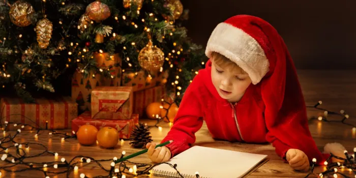 christmas child boy writing letter to santa kid in xmas hat writing gift wish list next to decorated christmas tree over dark background looking and lying down