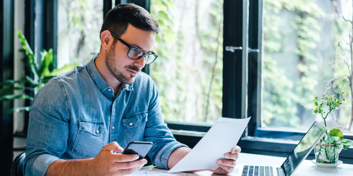 serious pensive thoughtful focused young casual entrepreneur small business owner accountant bookkeeper in office looking at and working with laptop and income tax return papers and documents
