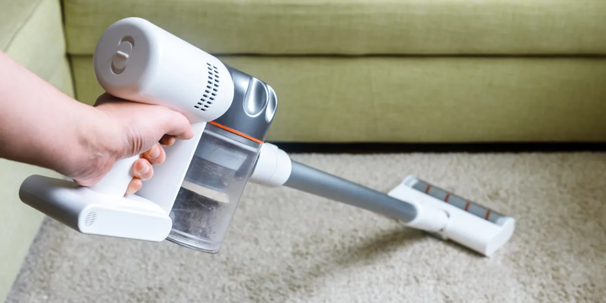 wireless vacuum cleaner used on carpet in room housework with new upright hoover person holds modern white vacuum cleaner by sofa home cleaning, care and technology concept