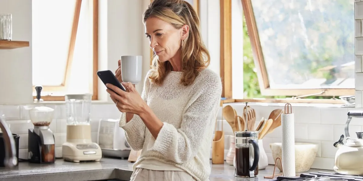 coffee, phone and senior woman in kitchen browsing social media, text message or online app at home relax, tea and reading news or mobile online web surfing with female from canada on 5g smartphone
