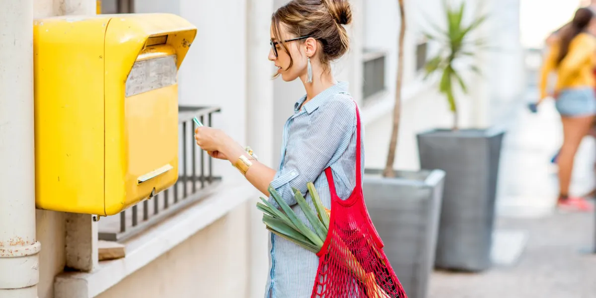 young woman putting letter to the old yellow mailbox standing with mesh bag full of food outdoors on the street