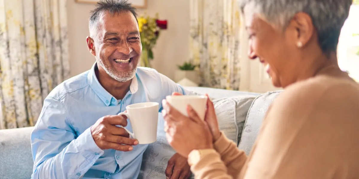 coffee, laughing and a senior couple on a sofa in the home living room to relax while bonding in conversation smile, retirement or love with a mature married man and woman talking while drinking tea