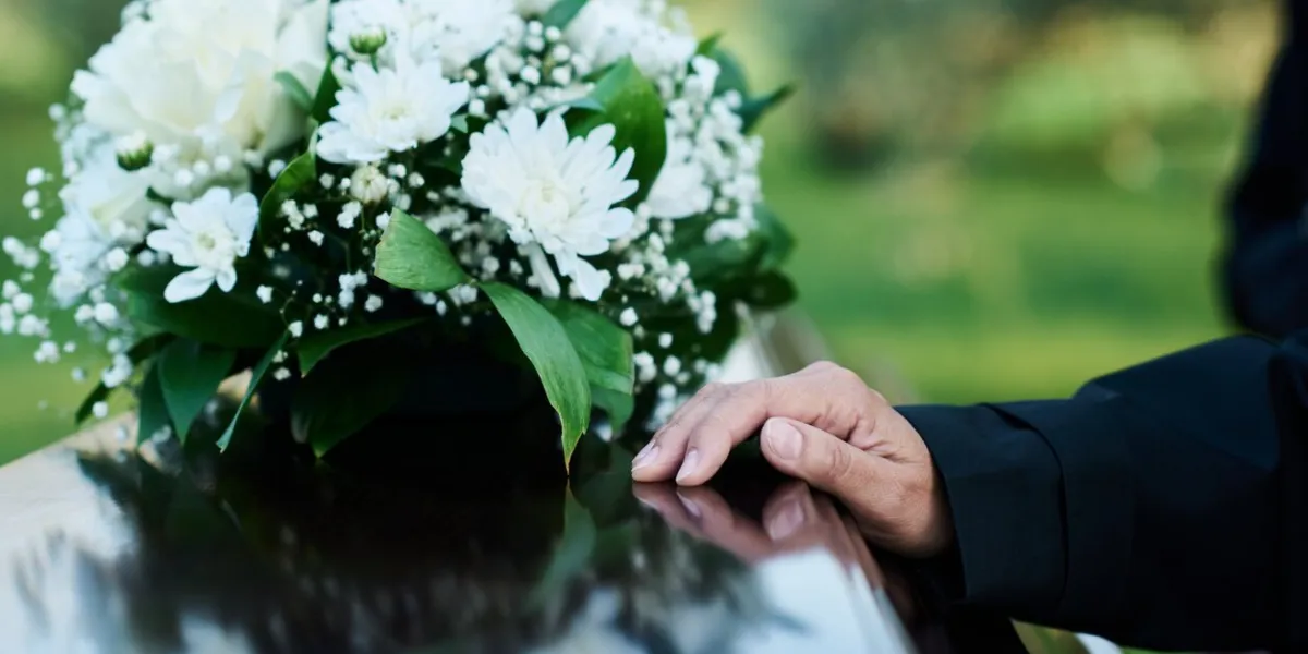 focus on hand of mourning mature woman in black attire on lid of closed wooden coffin with bunch of fresh white chrysanthemums on top