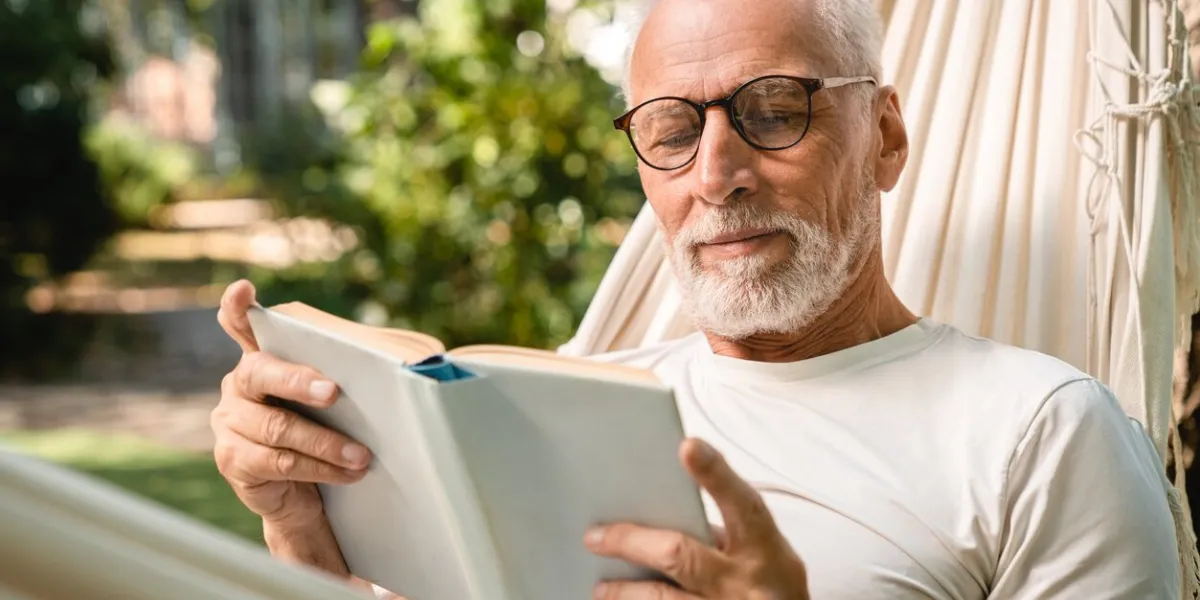 concentrated dreamy old senior elderly caucasian grandfather man relaxing resting in hammock while reading book outdoors in park garden forest