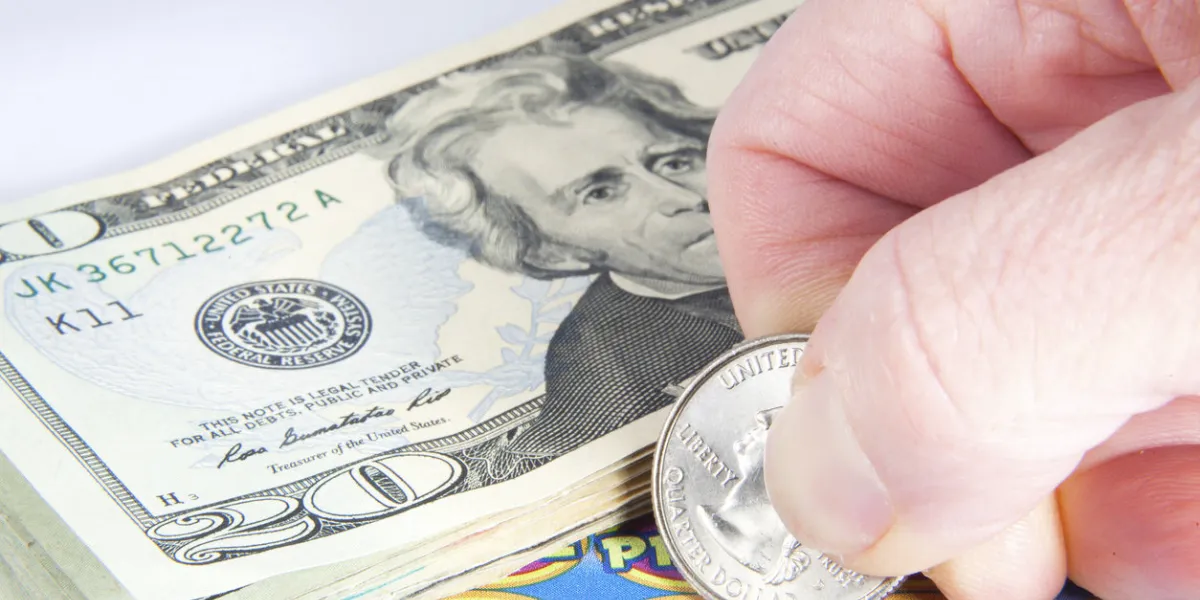 hand holding a quarter on a scratcher ticket with a pile of bills on top