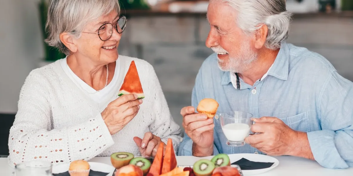 cheerful beautiful senior couple at home having breakfast together with muffin, milk and fresh seasonal fruit, healthy eating concept