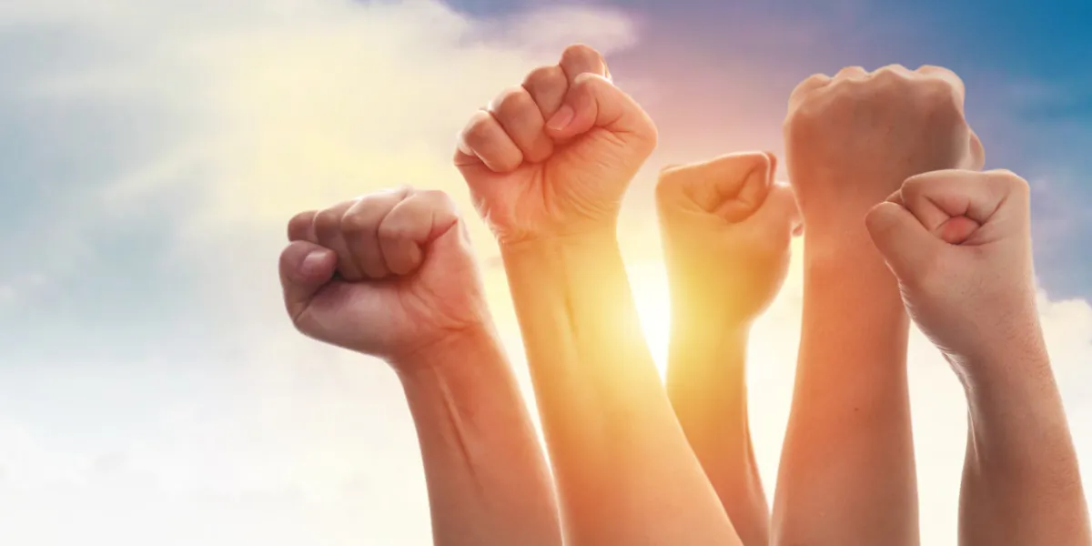 rising fist of adult people, male and female, over dramatic blue sky with sun light, anger protest revolution inspiration motivation or teamwork concept