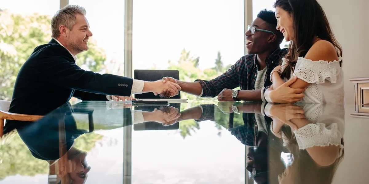 male real estate broker shaking hands with new property owners while sitting across a table property seller congratulating couple on making deal on new house