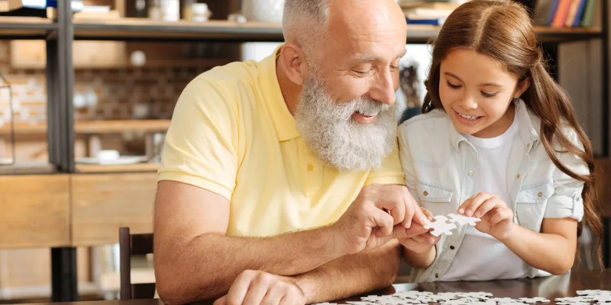 weekend in harmony adorable little girl and her pleasant grandfather sitting at the table, holding pieces of a jigsaw puzzle and trying to connect them together while smiling serenely