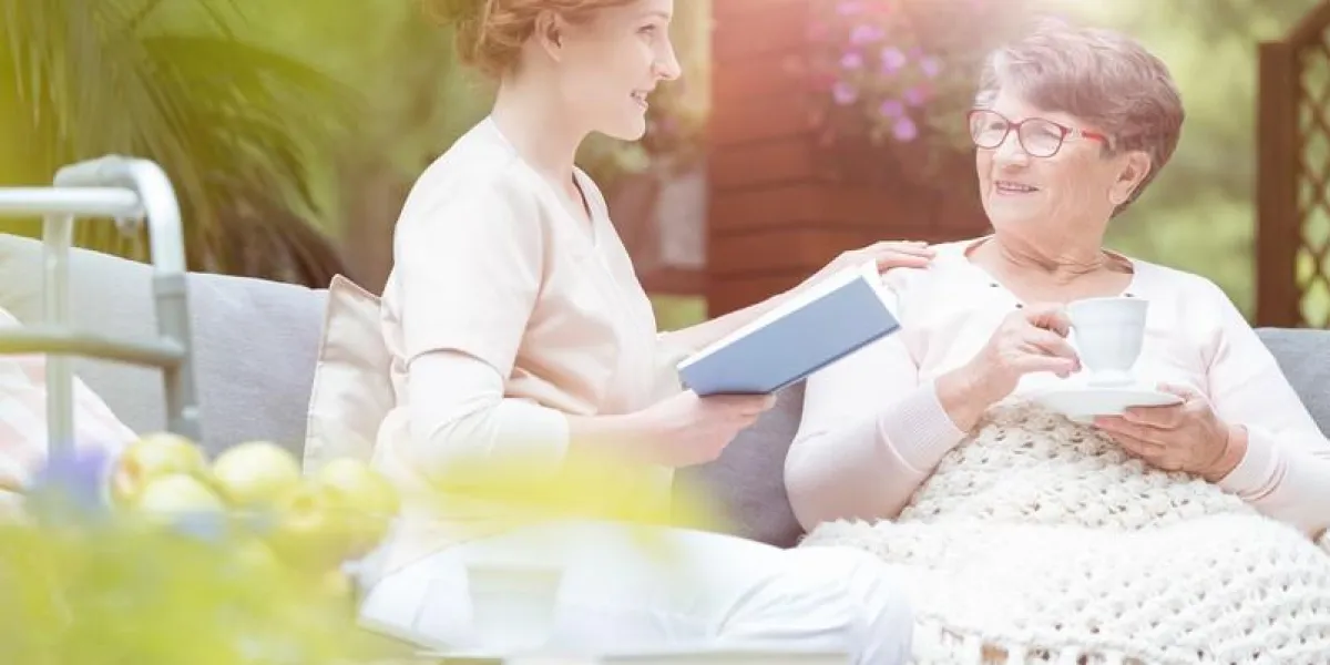 old woman talking with young caregiver reading book during meeting on terrace
