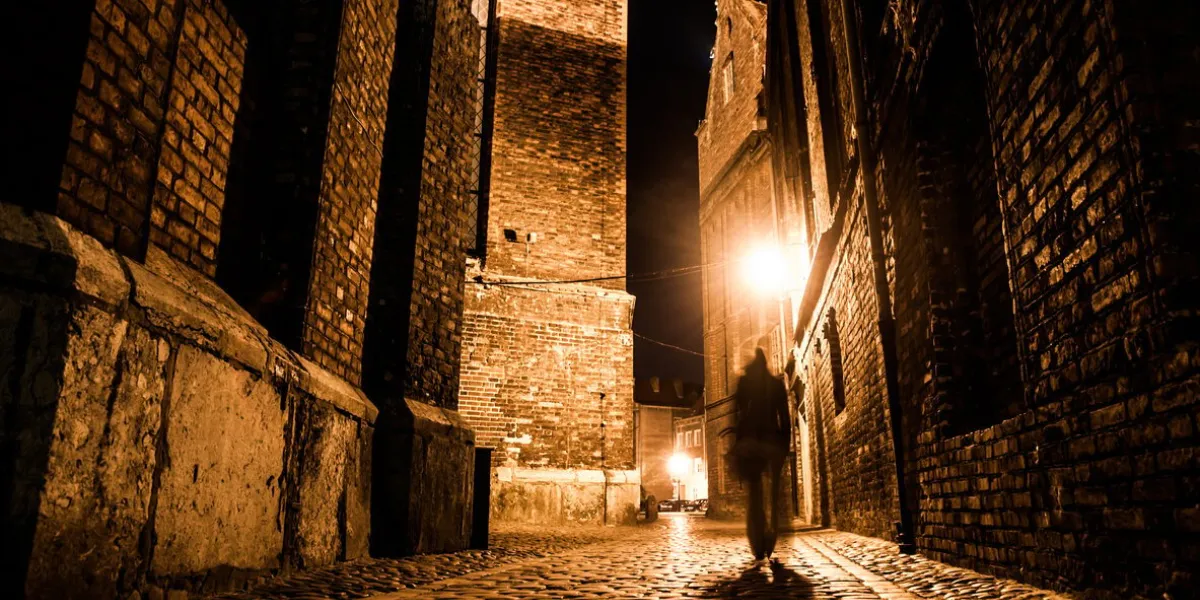 illuminated cobbled street with light reflections on cobblestones in old historical city by night dark blurred silhouette of person evokes jack the ripper