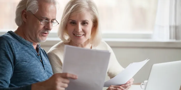 happy old middle aged couple holding reading good news in document, smiling senior mature family excited by mail letter, checking paying domestic bills online on laptop, discussing budget planning