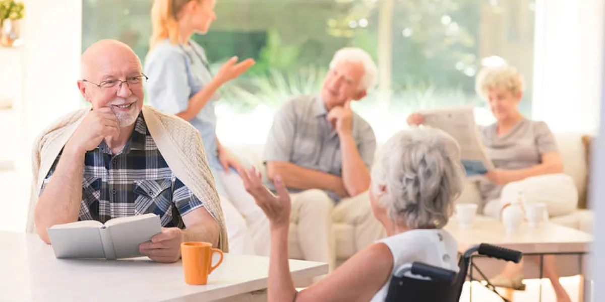 elderly man talking with disabled woman while sitting together at table in common room