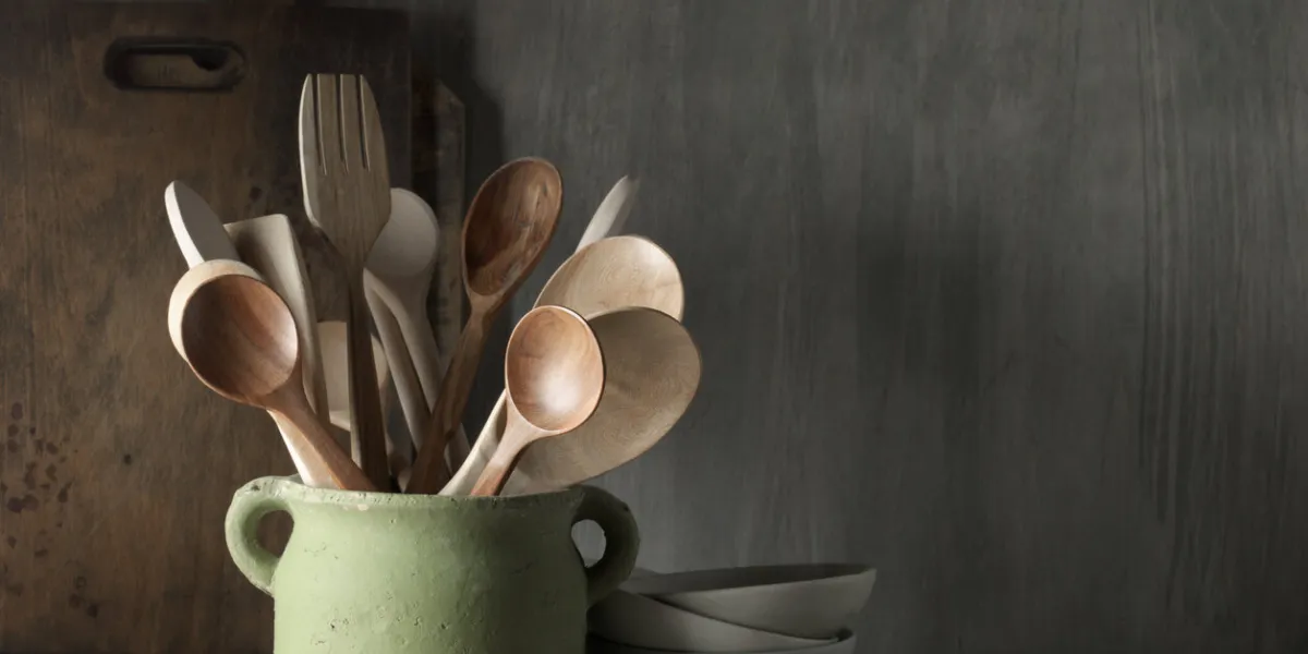 kitchen utensil set in rough handmade ceramic pot, crockery, cutting boards on wood table against rustic wooden wall low light