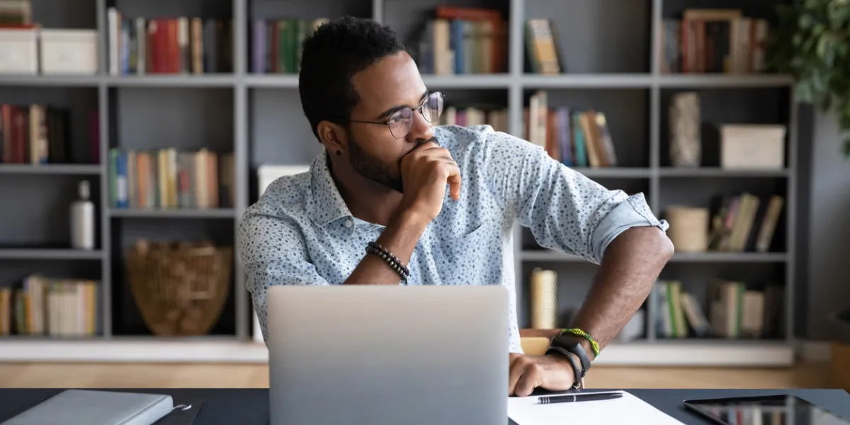 thoughtful doubtful african businessman in tension sitting at workplace desk near laptop distracted from work makes difficult decision looking at window feels uncertain thinking over problem solution