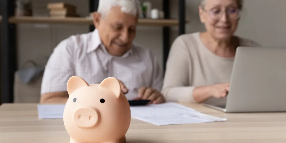 focus on piggybank on table, on background frugal elderly couple make payment using e-bank on laptop, calculates earning smile feel happy family budget control, management, investment, profit concept