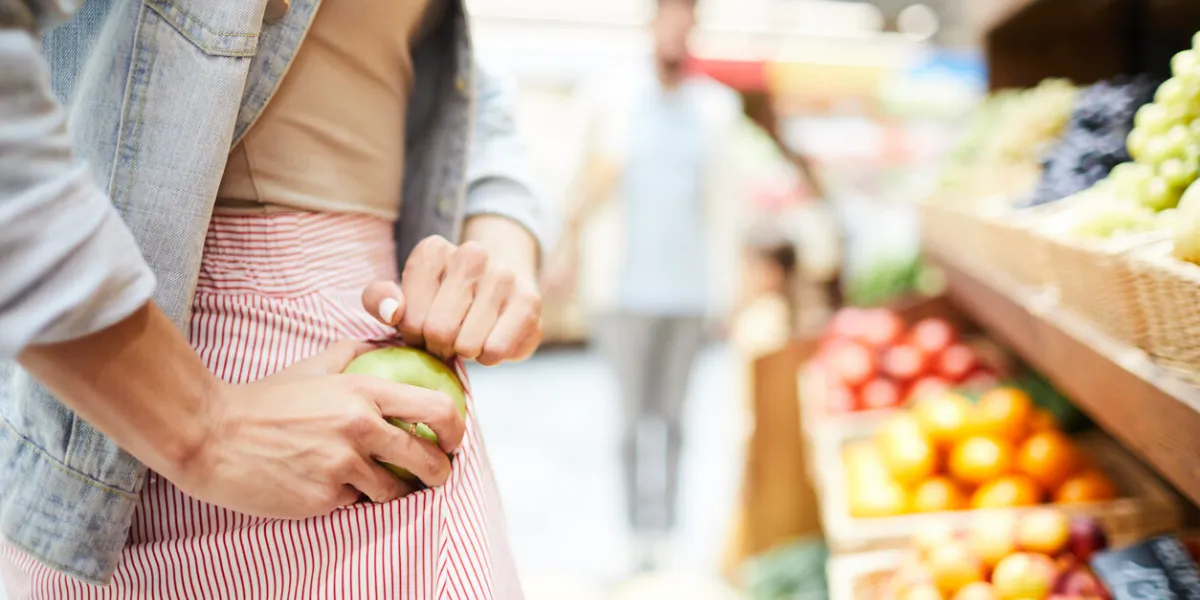 close-up of unrecognizable woman in stripped skirt hiding apple in pocket while stealing it in food store