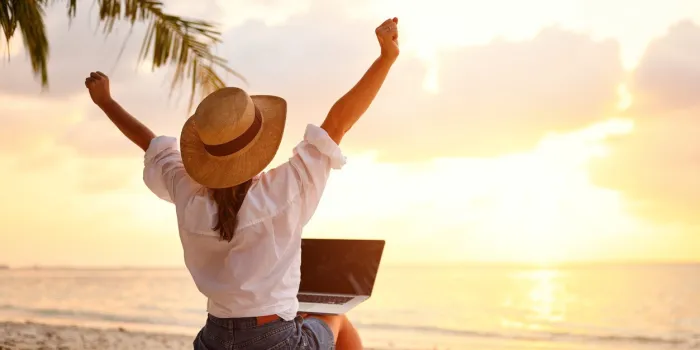 work from anywhere rear view of young woman, female freelancer in straw hat working on laptop, keeping arms raised and cellebrating success while sitting on the tropical sandy beach at sunset