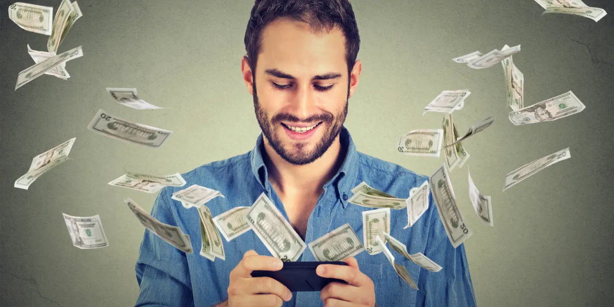 technology online banking money transfer, e-commerce concept happy young man using smartphone with dollar bills flying away from screen isolated on gray wall office background