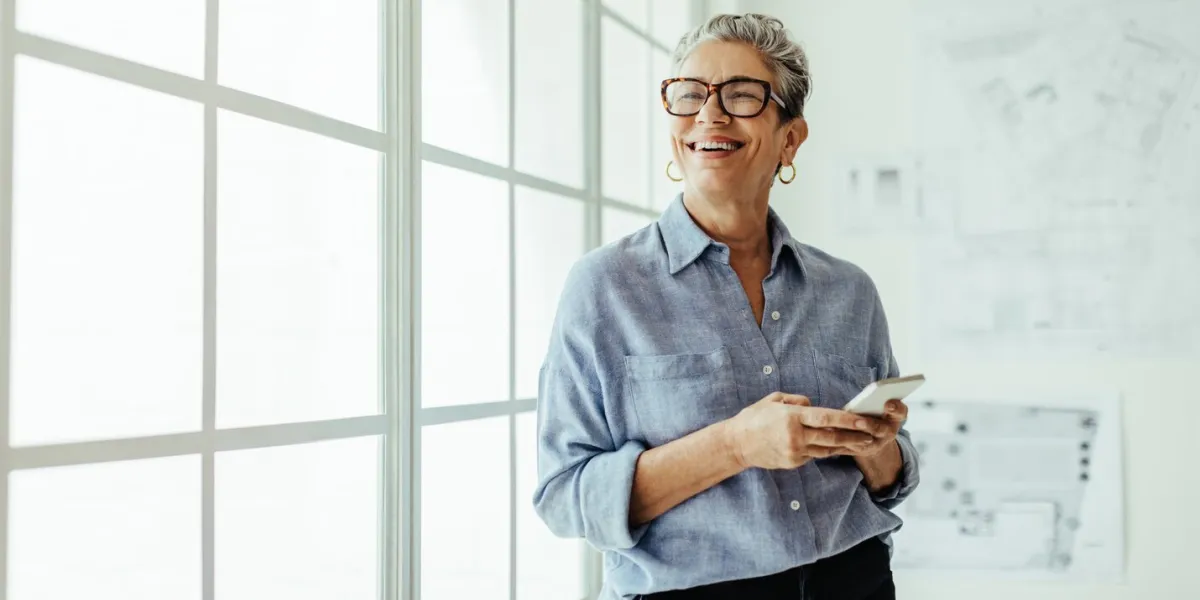 mature business woman stands in an office, holding a smartphone and looking outside the window with a smile female designer using mobile technology to stay connected with her colleagues and clients