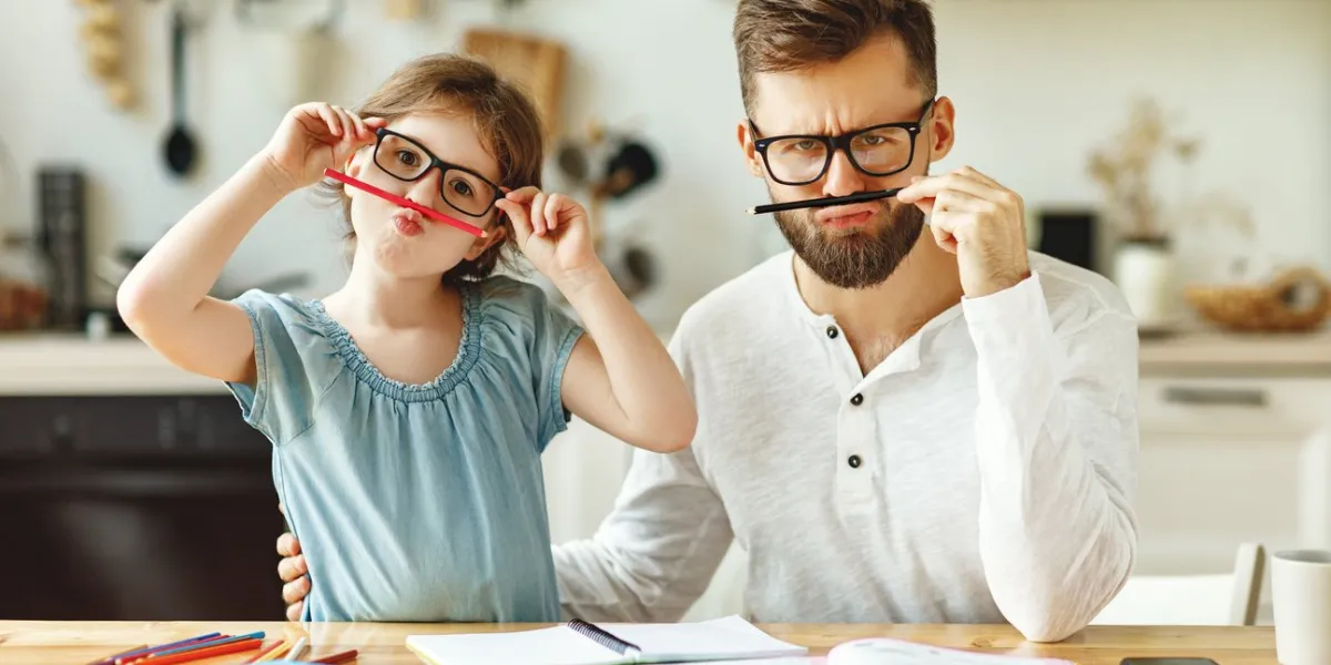 cheerful schoolgirl with dad wearing similar eyeglasses having fun and looking at camera while resting during studies at home