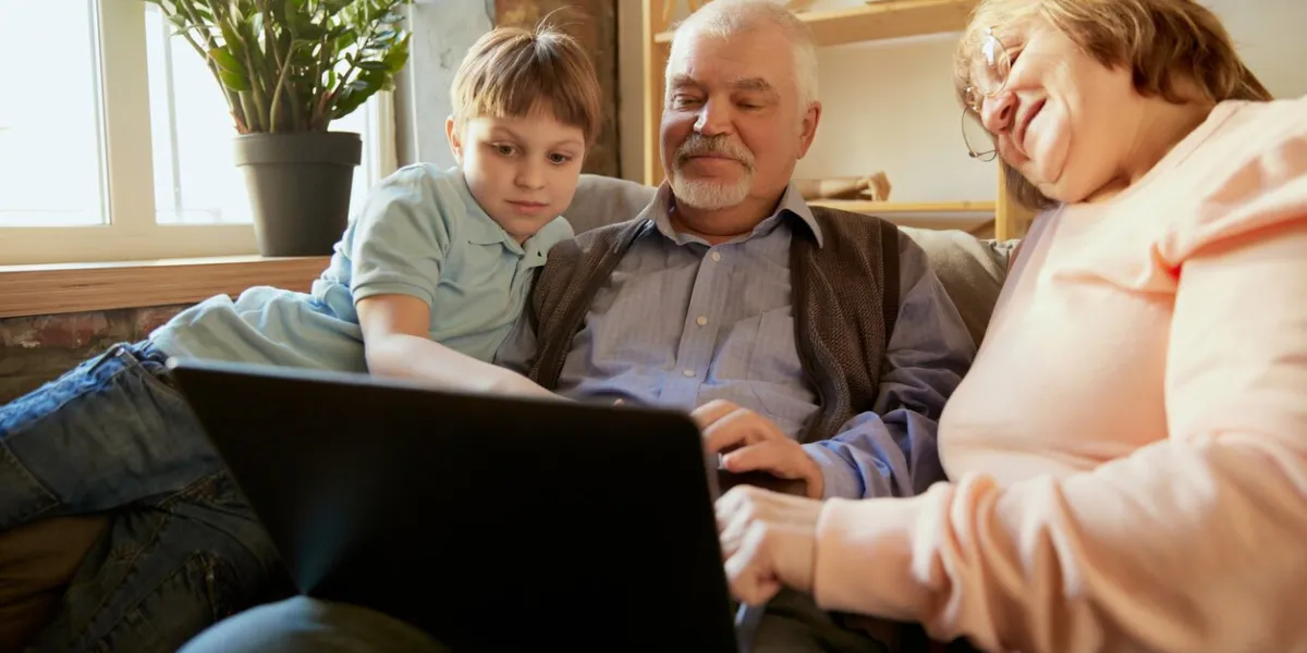life portrait of friendly family, grandparent and their grandson sitting on sofa and spending time together, using modern gadgets, talking, studying concept of love, care, support, hobbies and leisure activities