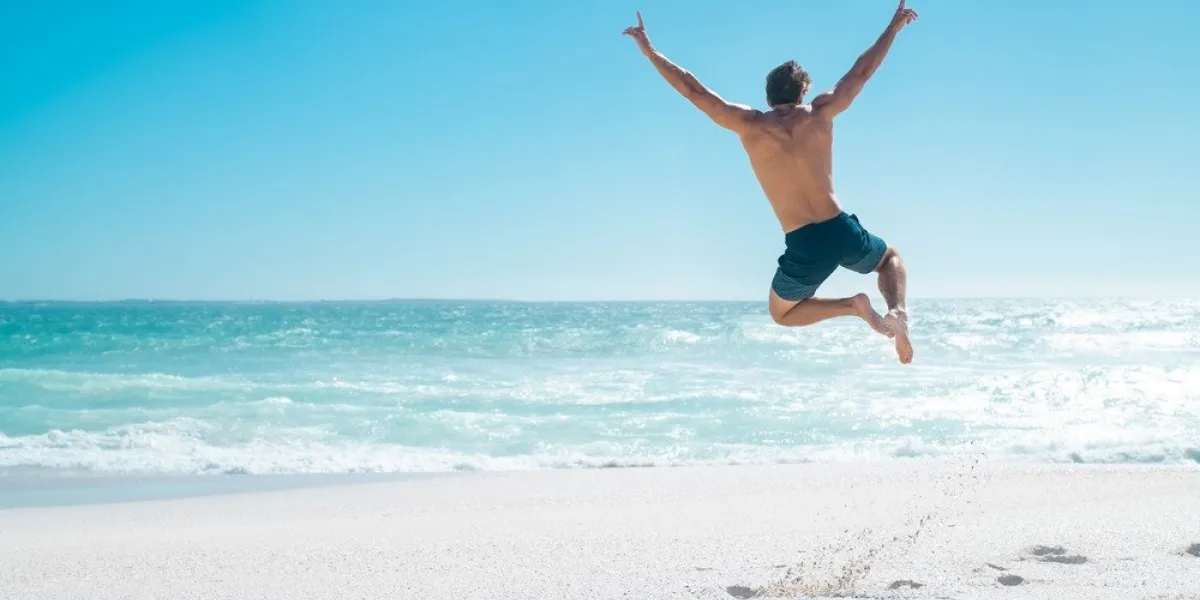 back view of happy young man jumping with outstretched arms on the beach with copy space rear view of shirtless guy wearing swimwear and having fun on tropical beach during vacation carefree and freedom concept