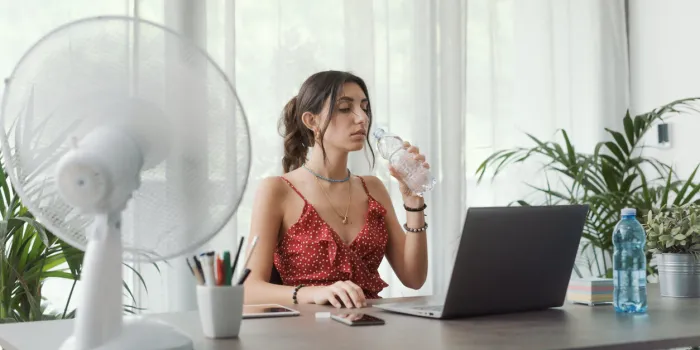 woman sitting at desk at home and working with a computer, she is cooling herself with an electric fan and drinking water
