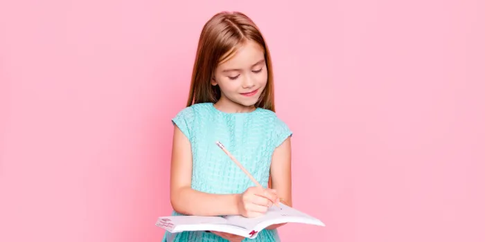 lovely adorable charming little girl is looking down at the copybook in her hands and writing information there, she is wearing light blue dress, isolated on bright pink background, copyspace