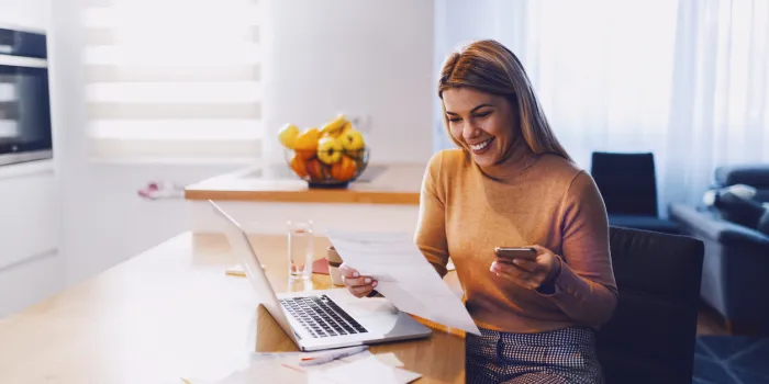 cute caucasian smiling blonde woman in sweater holding bills in one hand and in other smart phone on table are laptop and bills apartment interior