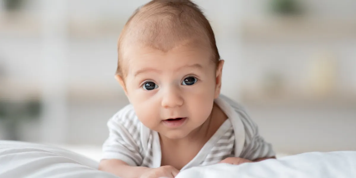 closeup portrait of cute little infant child lying on his belly on bed at home, adorable newborn baby looking at camera and learning how to crawl while relaxing on white linens, free space