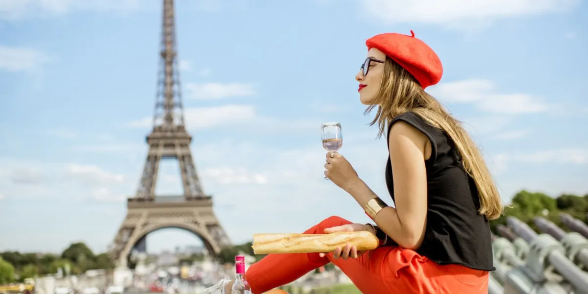 young woman in red cap having a picnic with glass of wine and baguette sitting in front of the eiffel tower in paris