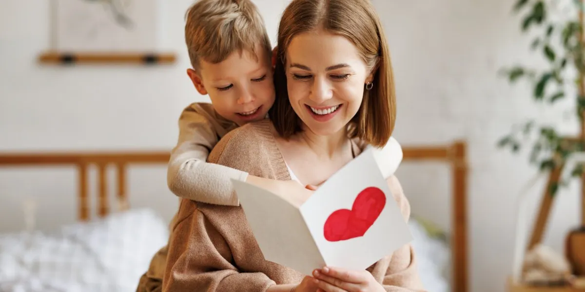 cute little boy son congratulating his mom happy woman with mothers day, giving her handmade greeting postcard with red heart while sitting together on bed at home family holidays concept